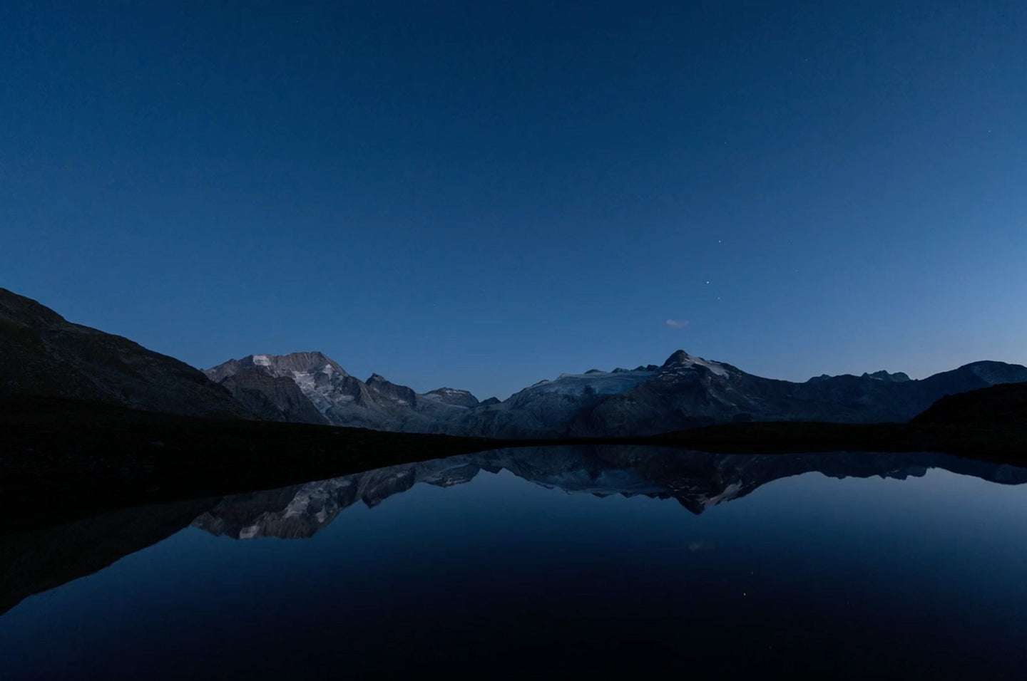 A serene mountain landscape at dusk, with snow-capped peaks reflecting on a still, glass-like lake under a deep blue sky with faint stars. The rugged slopes frame the tranquil water, creating a peaceful and symmetrical scene.