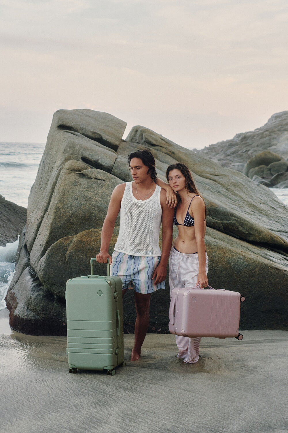 Two travelers stand barefoot at the shoreline with the Hybrid Trunk in Mint and the Hybrid Carry-On in Orchid, surrounded by ocean waves and coastal rock formations.