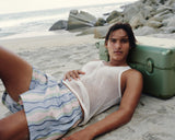 A man rests on the sand, leaning against the Hybrid Trunk in Mint with the ocean and rocks in the background.