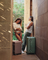 The Hybrid Carry-On in Orchid and Hybrid Trunk in Mint, captured in a quiet moment between two travelers in an architectural escape.