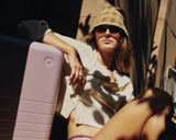 A woman lounges in the sun beside the Hybrid Carry-On in Orchid, dressed in soft neutrals and shadowed light.
