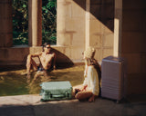 The Hybrid Carry-On in Mint and the Hybrid Trunk in Orchid beside two people relaxing by a sunlit pool framed in stone architecture.