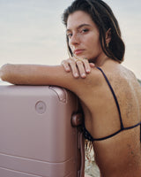 A woman with sand-dusted skin leans on the Hybrid Trunk in Orchid, gazing softly against a serene coastal backdrop.