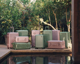 A full collection of Hybrid luggage in Mint and Orchid arranged by a tranquil pool, framed by lush greenery and stone architecture.