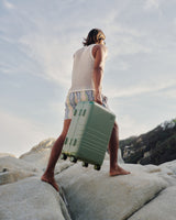 A barefoot man stands atop sunlit rocks, carrying the Hybrid Trunk in Mint against an open sky.