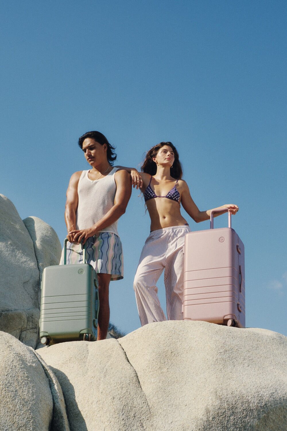 Two travelers stand on sunlit rocks with the Hybrid Carry-On in Mint and the Hybrid Trunk in Orchid, basking in the calm of a clear blue sky.