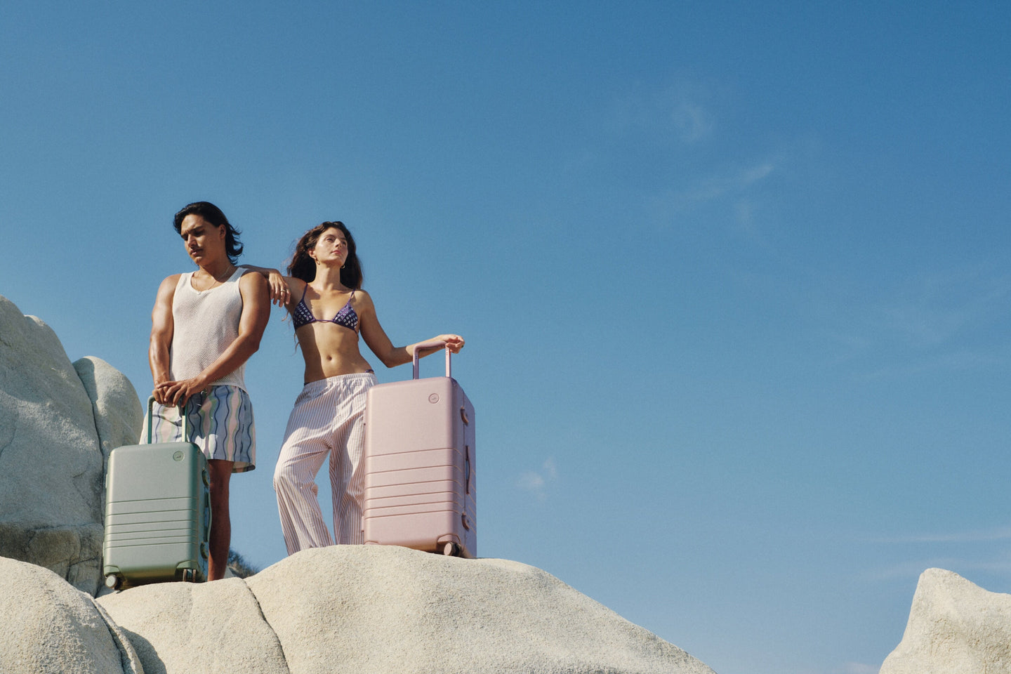 Two travelers stand on sunlit rocks with the Hybrid Carry-On in Mint and the Hybrid Trunk in Orchid, basking in the calm of a clear blue sky.